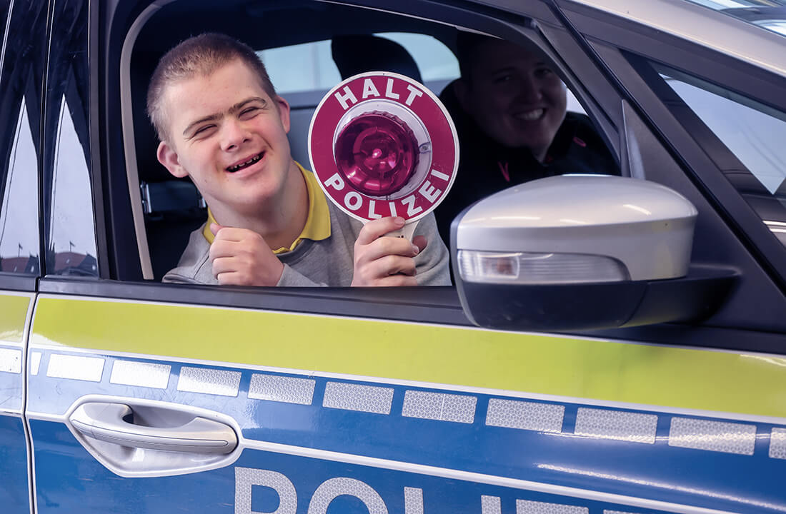Dominik sitzt in einem Einsatzwagen. Er hält eine rote Polizeikelle und lächelt..