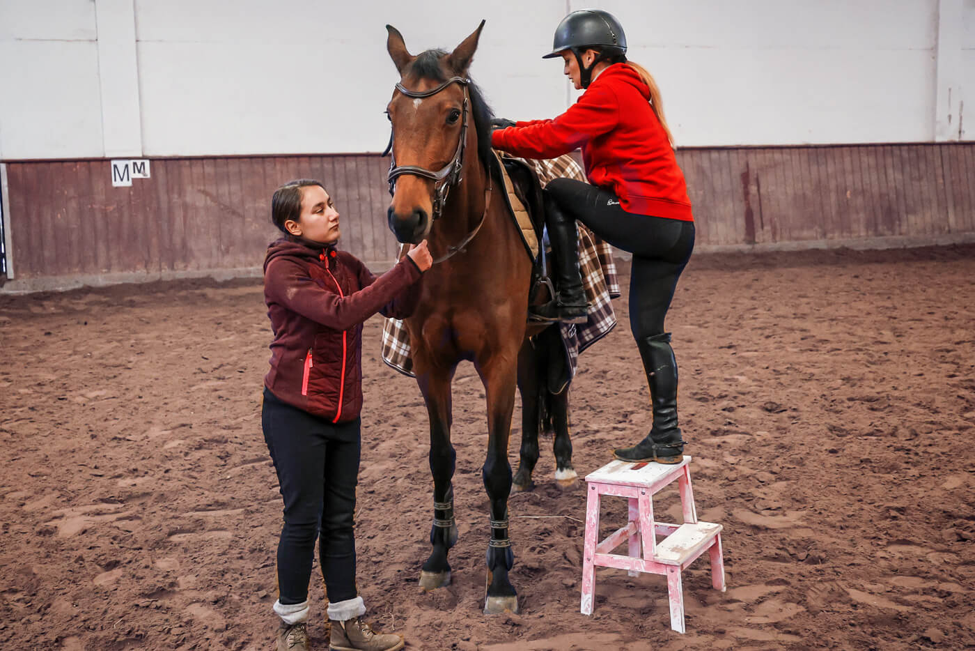 Eine zierliche Frau steigt von einer Trittleiter auf ein großes Reitpferd. Das Pferd wird von einem jungen Mädchen an der Trense festgehalten.