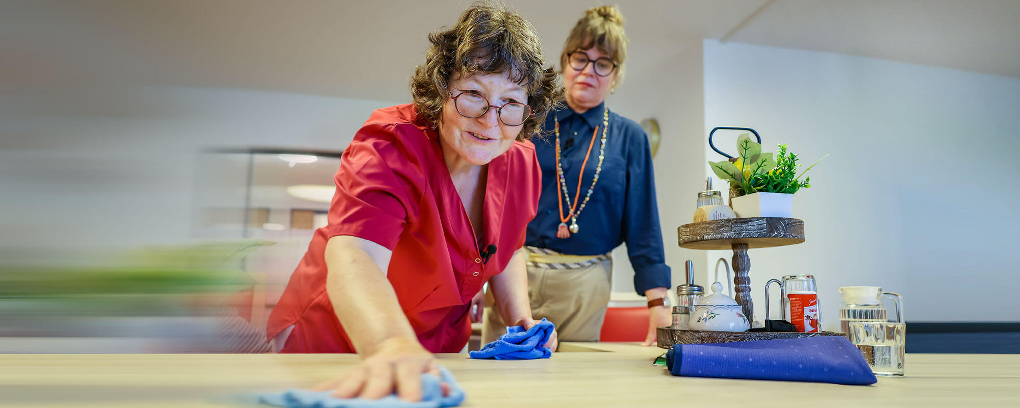 Eine Mitarbeiterin wischt einen Tisch. Ein Jobcoach schaut ihr dabei über die Schulter.