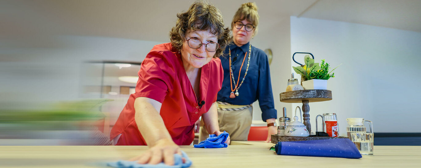 Eine Mitarbeiterin wischt einen Tisch. Ein Jobcoach schaut ihr dabei über die Schulter.