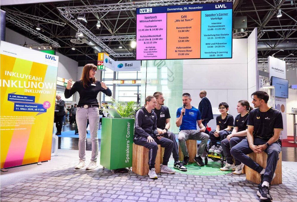 Eine Gruppe junger Menschen sitzt auf Holzblöcken am Messestand des LWL-Inklusionsamts Arbeit.