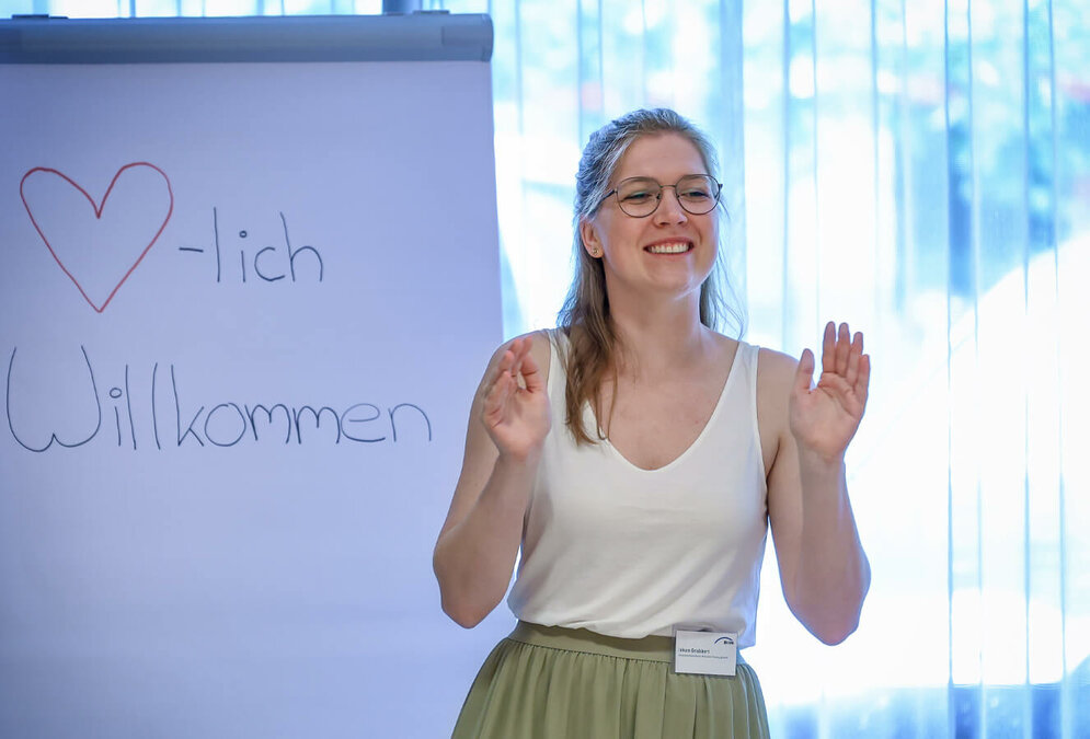 Eine junge Frau steht lächelnd vor einem Flipchart mit der Aufschrift "Herzlich Willkommen".