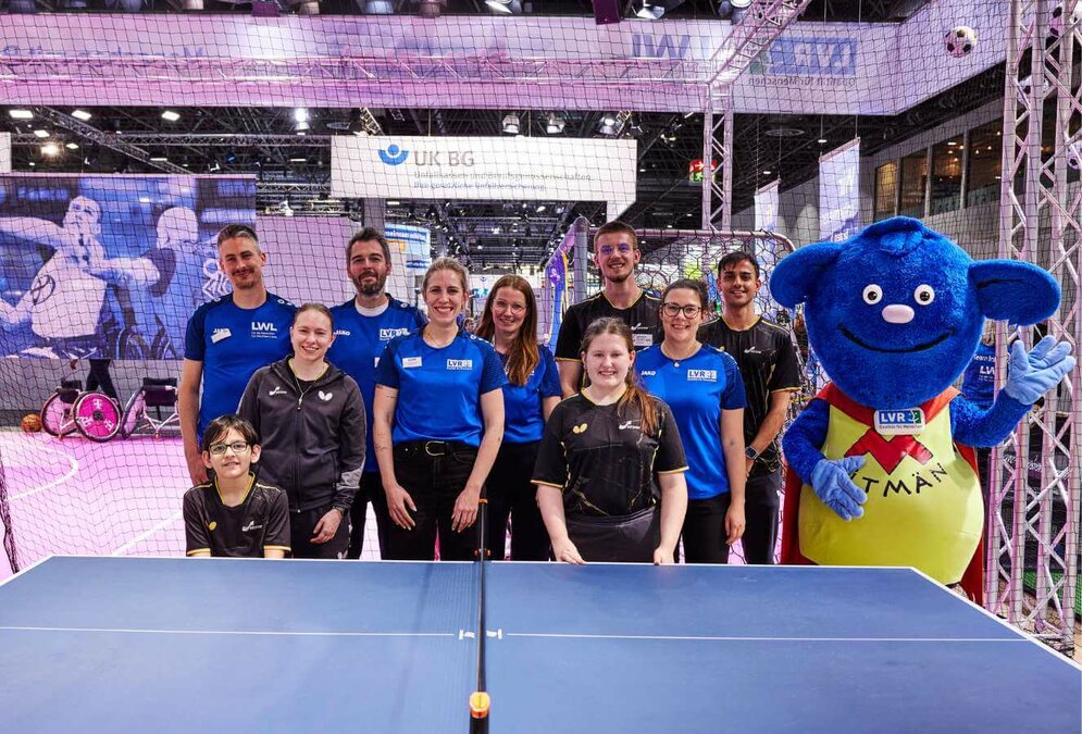 Eine Gruppe Mädchen und Jungen in Sportkleidung steht hinter einer Tischtennisplatte. Neben ihnen steht eine lebensgroße Plüschpuppe im Supermann-Kostüm. Auf dem gelben Shirt steht "LVR-Mitmän".
