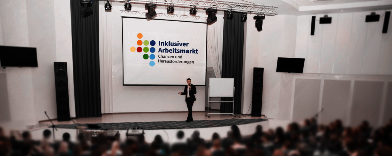 Das Foto zeigt einen großen, vollbesetzten Veranstaltungsraum. Auf der Leinwand im Bühnenbereich ist das Logo des Forums mit der Beschriftung eingeblendet: Inklusiver Arbeitsmarkt, Chancen und Herausforderungen