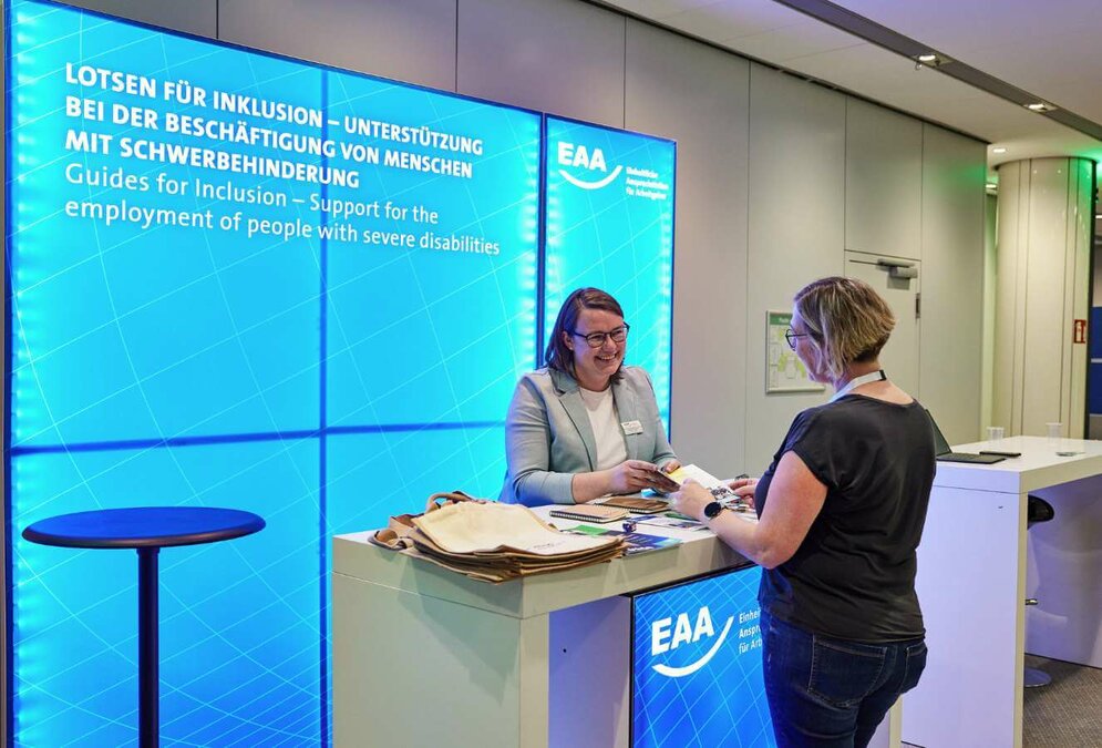 An einem Messestand stehen zwei Frauen und unterhalten sich. Auf der blauen Messewand ist zu lesen: "Lotsen für Inklusion – Unterstützung bei der Beschäftigung von Menschen mit Schwerbehinderung"