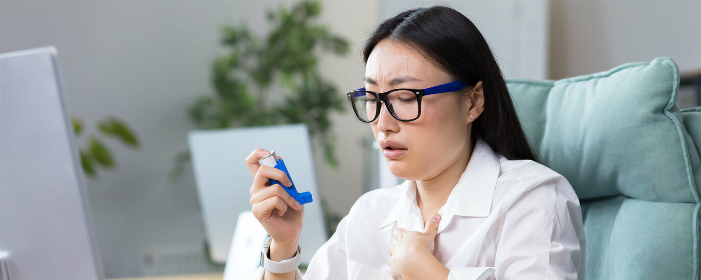 Foto einer jungen Frau mit dunklem Haar und Brille an einem Computerarbeitsplatz. Sie hält ein Inhalierspray in der rechten Hand und greift mit der linke an ihre Brust. Sie hat Probleme mit dem Atmen