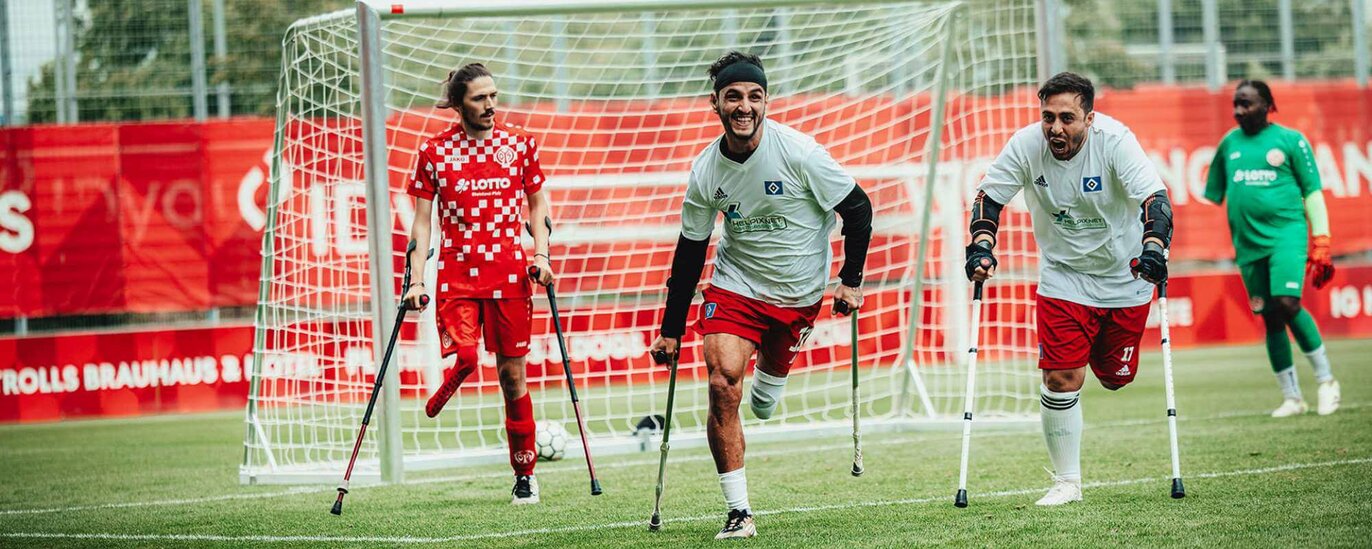 Das Foto zeigt drei beinamputierte DAFL-Fußballspieler vor dem Tor. Auf Unterarmstützen rennen sie aus voller Kraft dem Ball entgegen. Der Linienrichter ist armamputiert.