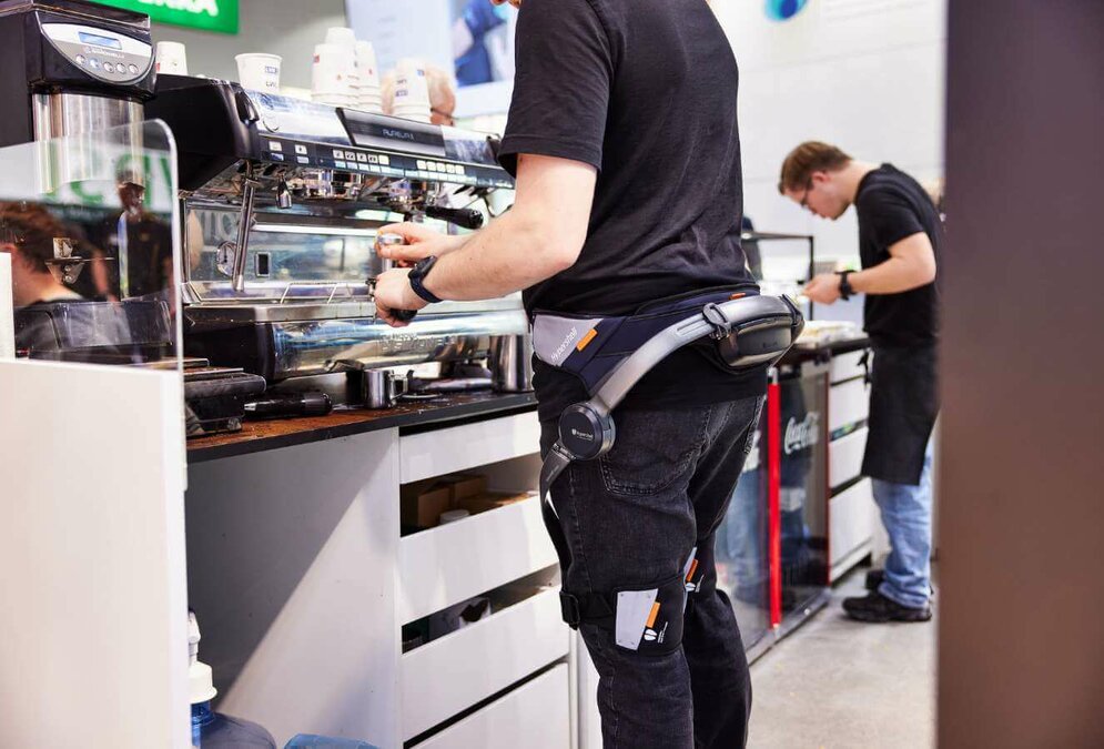 Zwei junge Männer, einer davon mit Down-Syndrom, stehen an einer Café-Bar. Vom vorderen Mann sind nur die Beine und ein Teil des Oberkörpers zu sehen. Er trägt Exoskelett, das Hüfte und Beine stützt. 