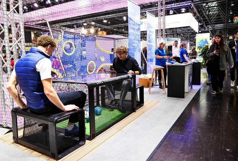 In einer großen Messehalle ist vorne links im Bild ein länglicher Tisch zu sehen unter dem es ein mit Netzen geschütztes Fußballfeld gibt. Zwei Jungen sitzen am Kopf- und Fußende des Tisches und kicken einen kleinen blauen Ball hin und her.