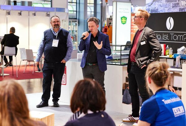 Drei Männer stehen auf einer Messe vor Publikum. Der mittlere Mann hält ein Mikrofon. Die beiden anderen schauen ihn an.