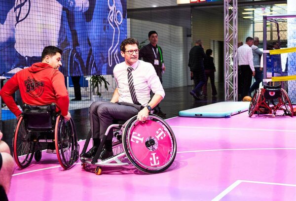 Auf einem pinkfarbenem Rollstuhlbasketballfeld fahren zwei Männer ganz dicht aufeinander zu. Der Mann rechts trägt eine Anzughose und ein weißes Hemd. Seine Krawatte weht leicht im Fahrtwind.