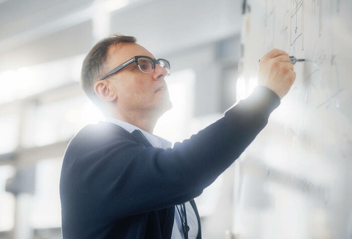 Ein Mann mittleren Alters mit Brille steht an einem Whiteboard und schreibt etwas auf die Tafel.