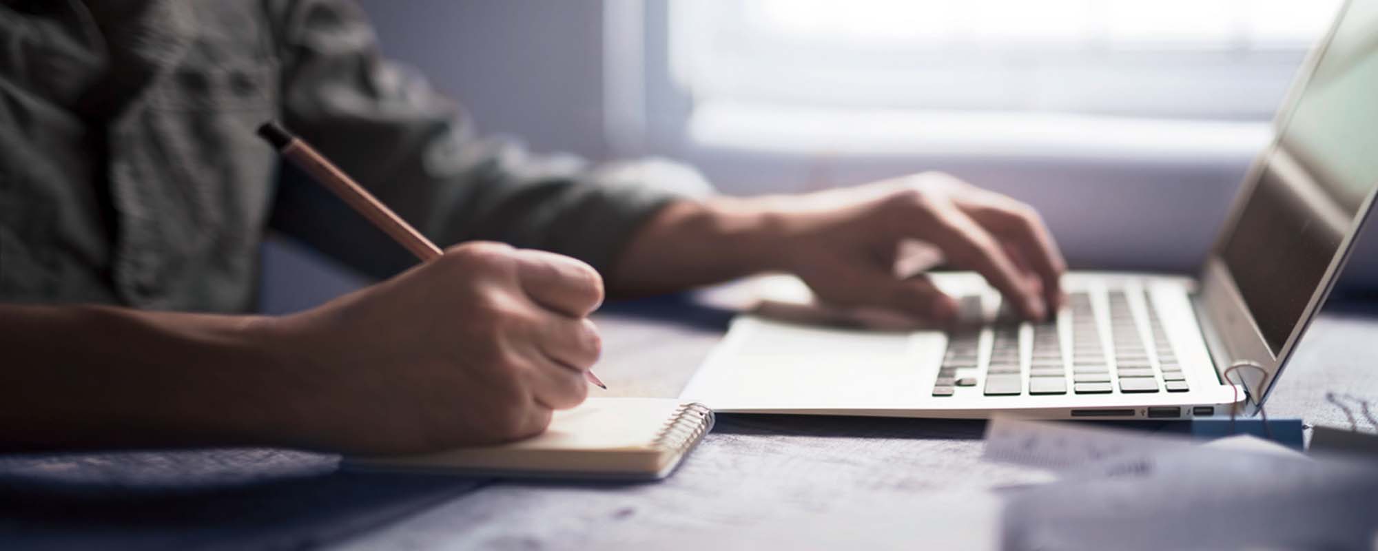 Mann am Schreibtisch | © shutterstock / Dhima Mann sitzt mit Laptop und Block und Stift am Schreibtisch.