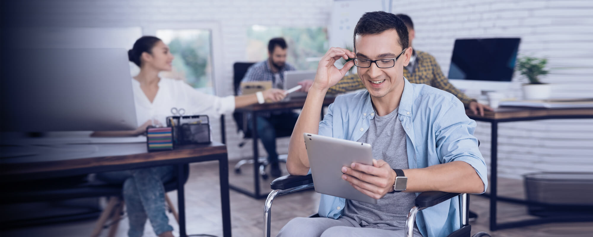 Ein junger Mann sitzt im Rollstuhl in einem Büro und schaut lächelnd auf ein Tablet.