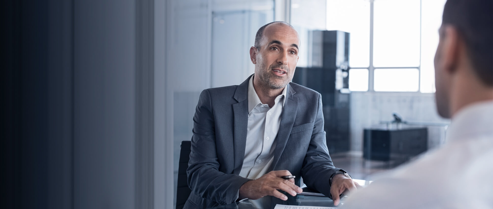 Mann in Anzug spricht in einem Büro mit einer weiteren Person| © shutterstock / Rido Mann in Anzug spricht in einem Büro mit einer weiteren Person.