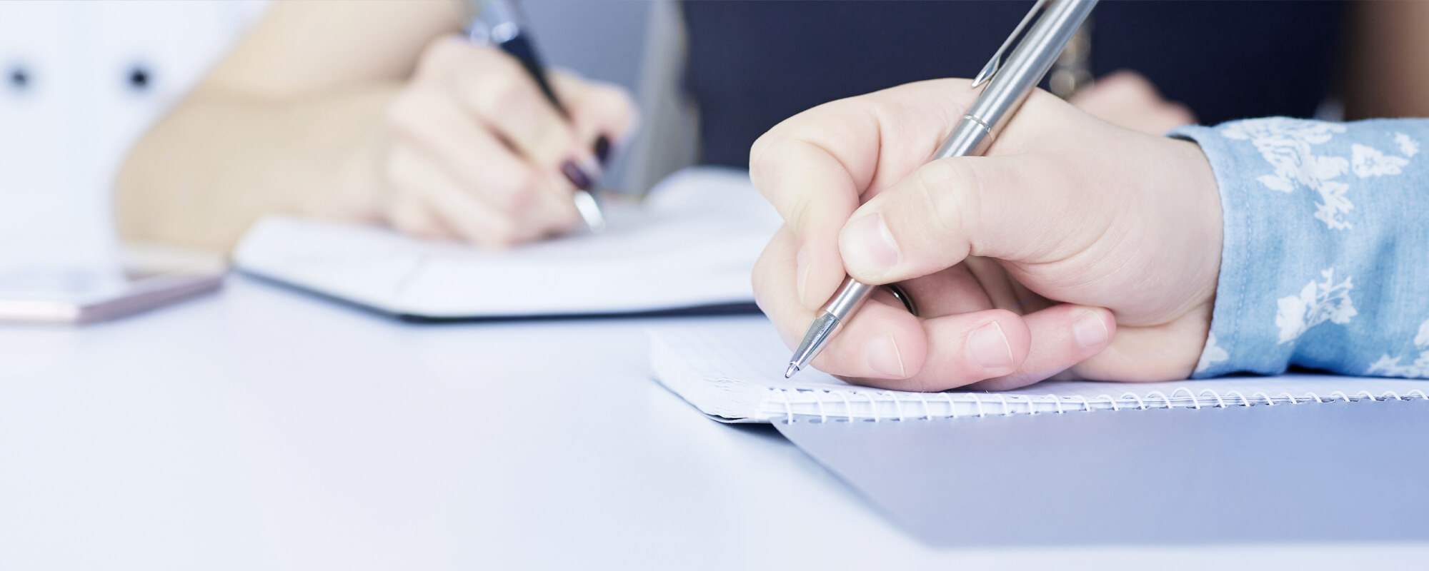 Personen mit Kugelschreiber | © shutterstock / Vitali Michkou Zwei Personen sitzen gemeinsam am Tisch und schreiben mit Kugelschreiber in Ringbücher.