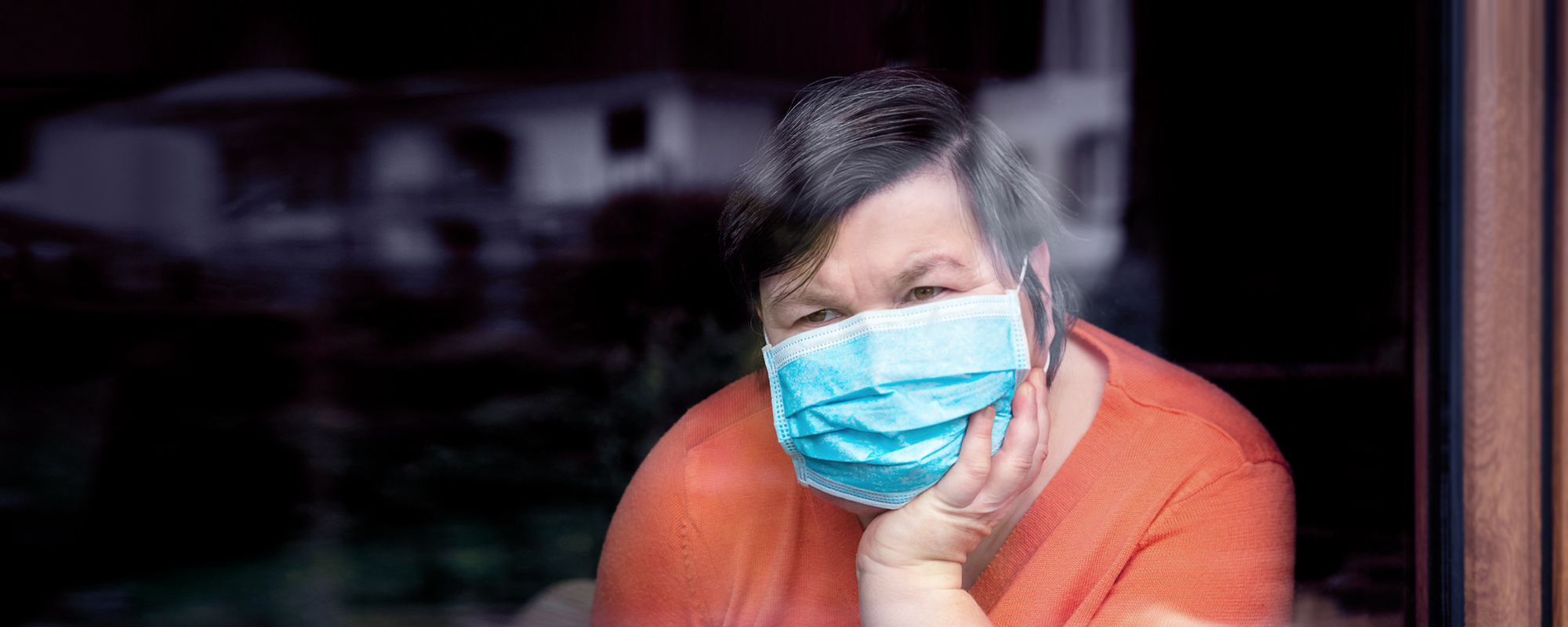 Ältere Frau mit medizinischem Mundschutz schaut nachdenklich aus einem Fenster. 