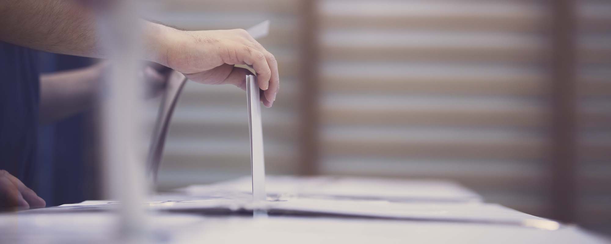 Person hält in der Hand einen Wahlzettel über dem Schlitz der Wahlurne. 