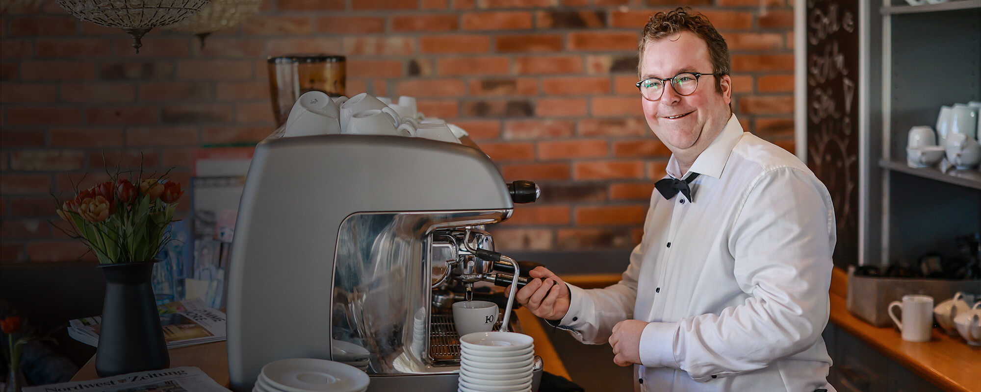 In einem Café bedient ein junger Mann mit Downsyndrom eine große Gastrokaffeemachine. Er trägt ein weißes Hemd und eine schwarze Fliege. Die Kamera blickt von der Seite auf ihn. Er lächelt in die Kamera.