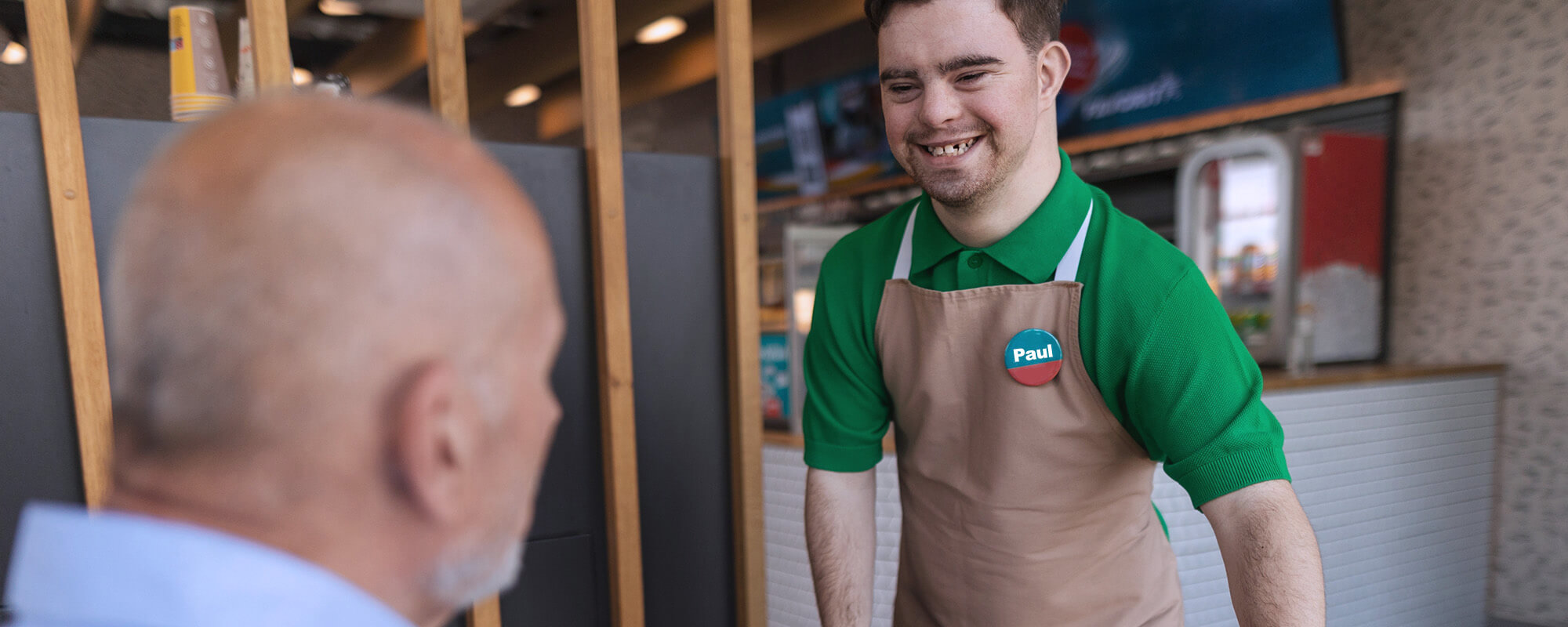 Das Foto zeigt einen jungen Mann mit Down-Syndrom. Er steht lächelnd an einer Ladentheke und bedient einen älteren Herren, von dem nur der Kopf im seitlichen Profil zu sehen ist.