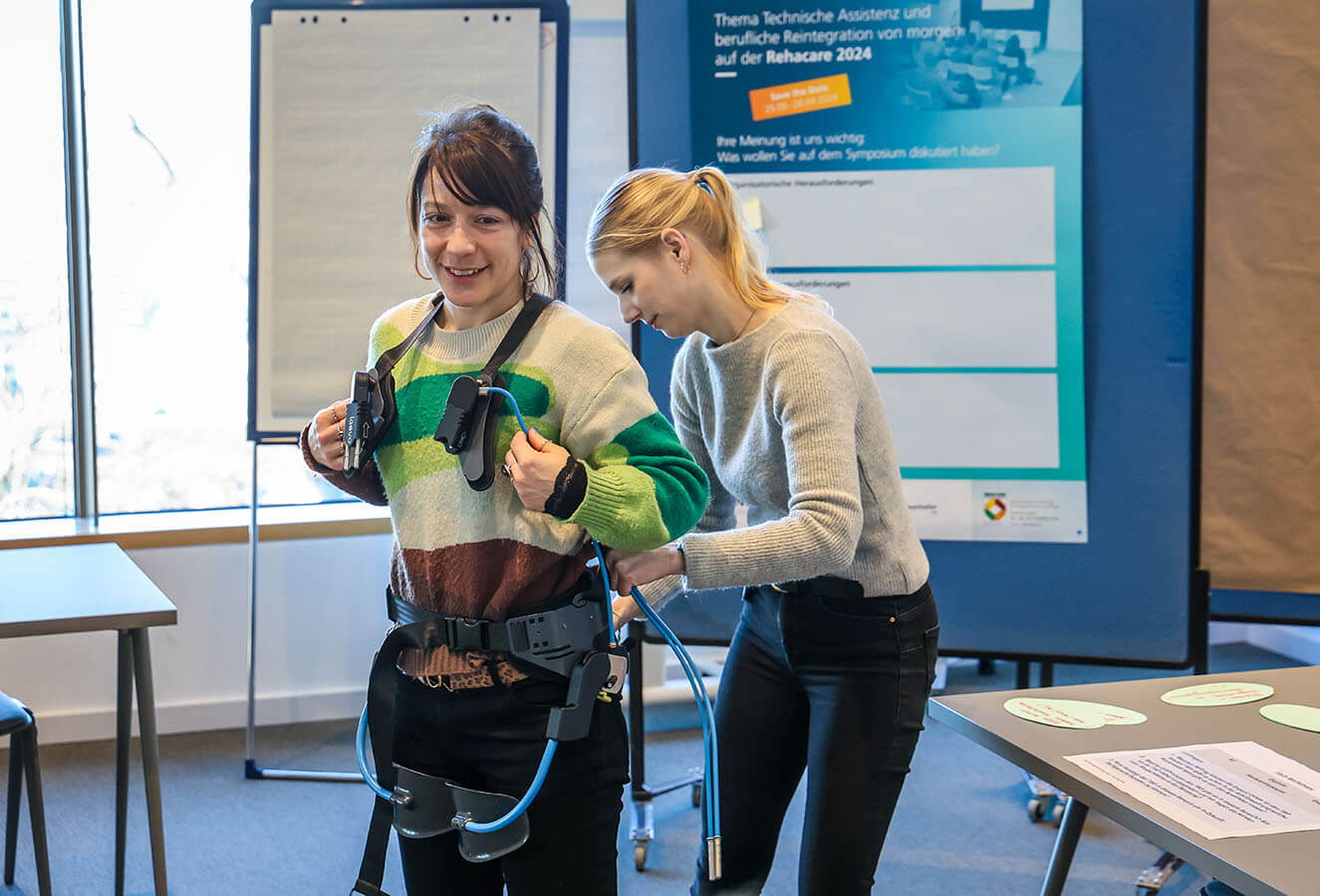 Das Foto zeigt zwei junge Frauen. Die Vordere trägt ein sogenanntes Exoskelett. Die Frau hinter hier, hilft beim Anziehen des Assistenzsystems.