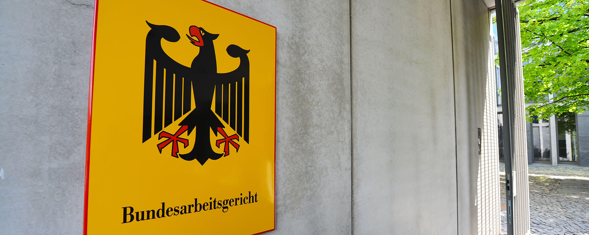 Das Foto zeigt ein gelbes Schild an einer großen Betonwand und Bäume im Hintergrund. Auf dem Schild steht "Bundesarbeitsgericht".