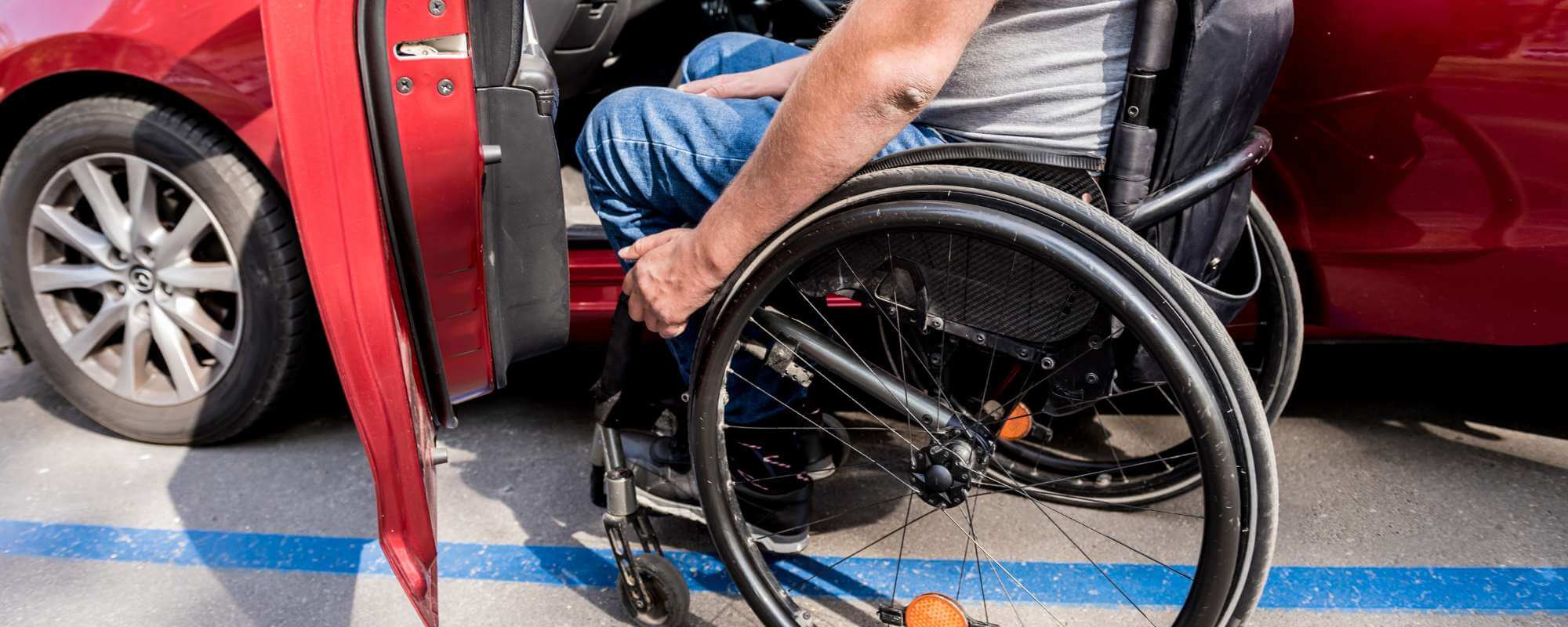 Auf der Fahrerseite eines roten Pkw steht Rollstuhl vor der geöffneten Tür des Fahrzeugs. Ein Mann mittleren Alters ist gerade dabei, vom Rollstuhl in das Fahrzeug hinüberzuwecheseln. 