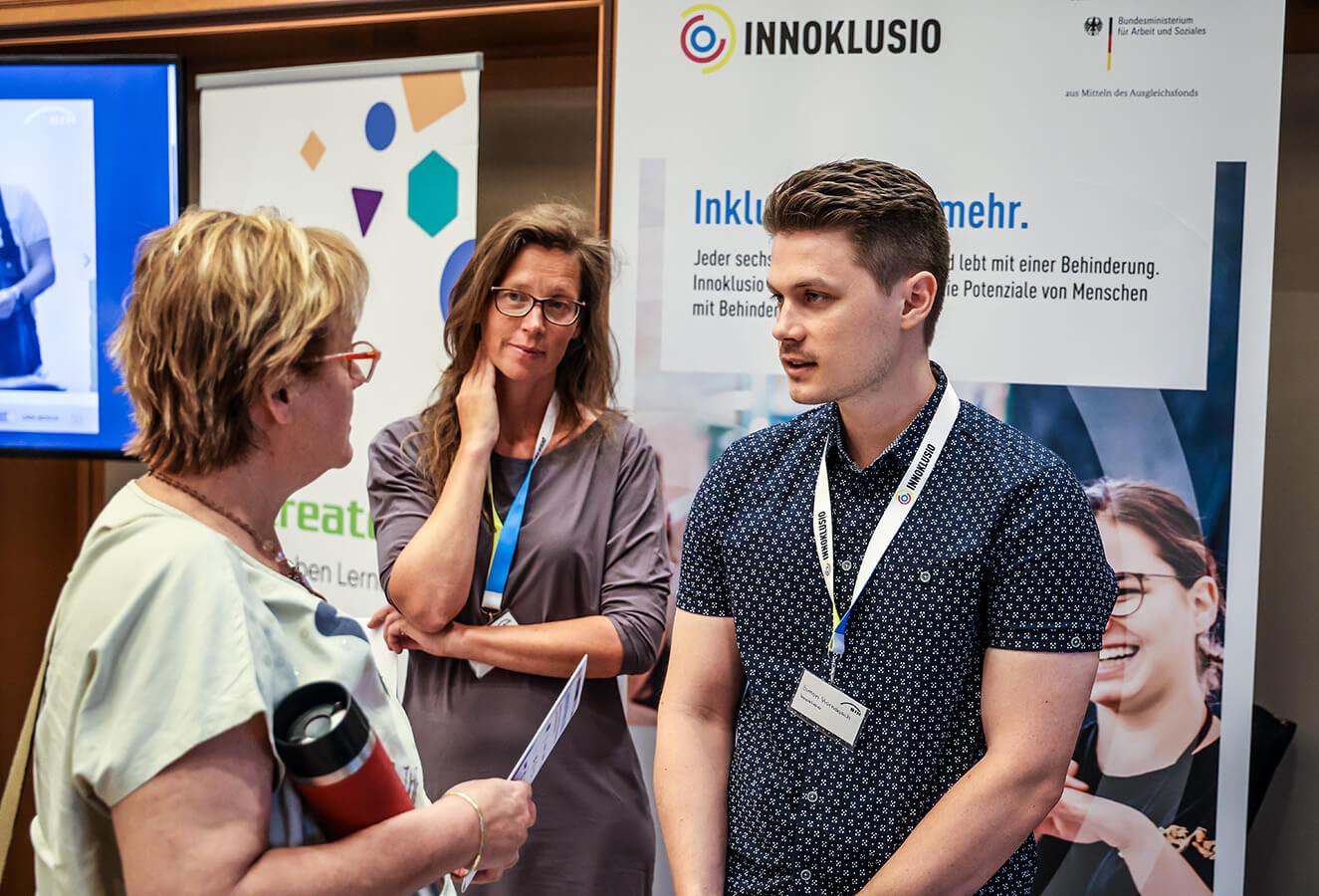 Eine Frau und ein junger Mann stehen am Infostand des Projekts Innoklusio. Eine weitere Frau lässt sich über Möglichkeiten der Zusammenarbeit von Menschen mit und ohne Behinderung beraten.