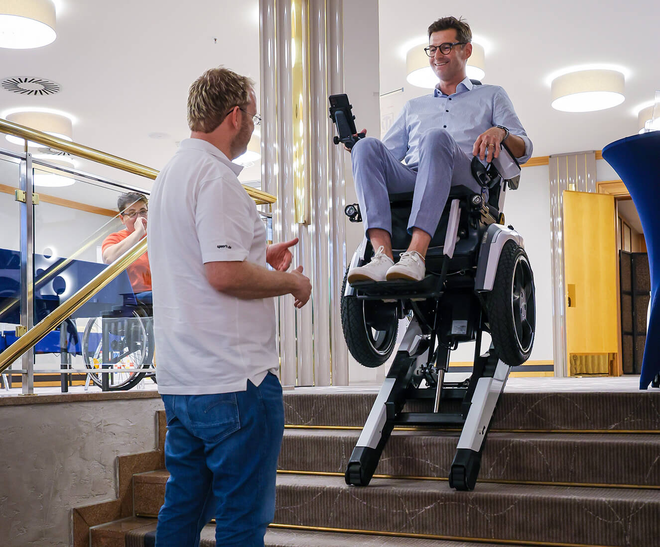 Ein Mann mittleren Alters fährt in einem Elektrorollstuhl mit Rädern und Raupen eine Treppe hinunter. Ein zweiter Mann am Fuß der Treppe gibt Anweisungen.