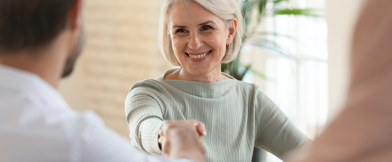 SBV-Antrittsbesuch beim Arbeitgeber | © VIACHESLAV/Shutterstock. No use without permission. Eine Frau zwischen 50 und 60 reicht einem jüngeren Mann die Hand und lächelt. Der Blick der Kamera fällt über die Schulter des Mannes auf die Frau.