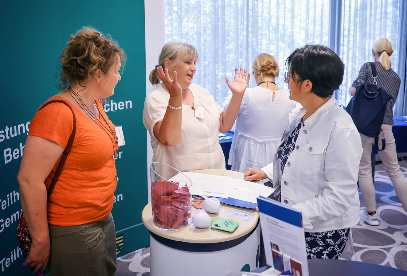 Drei Frauen stehen vor einem großen grünen Display an einem Stehtisch und unterhalten sich.