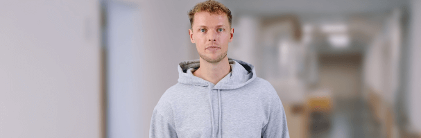 Das Foto zeigt einen Avatar, ein junger Mann im Hoody, der in einem langen Büroflur steht. Der Hintergrund ist etwas unscharf.