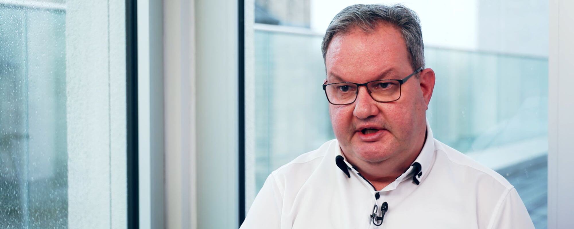 Das Foto zeigt eine Interviewsituation: Der Blick fällt von vorne auf den Oberkörper eines Mannes im mittleren Alter. Er trägt ein weißes Hemd ohne Krawatte.