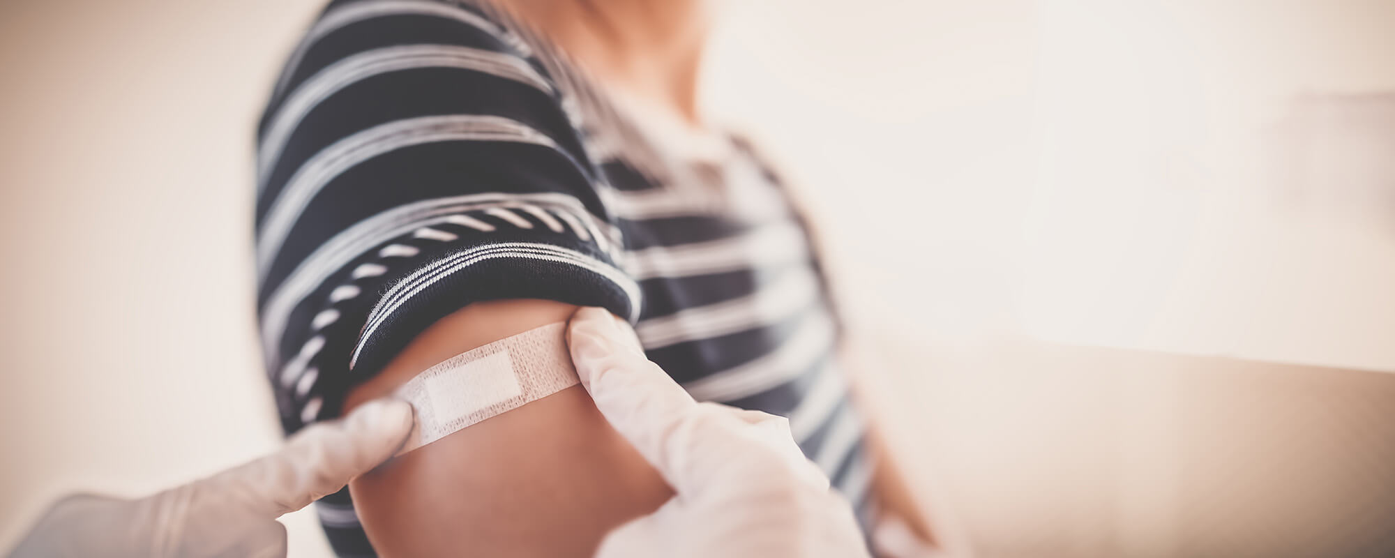 Zwei Hände in medizinischen Handschuhen kleben einer Person auf Pflaster auf den Oberarm. 