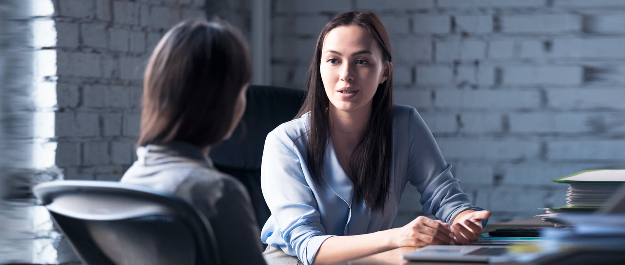 Zwei junge Frauen mit langen Haaren sitzen in einem Büro nebeneinander und reden miteinander.