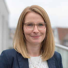 Foto von Simone Zimmer im Freien. Sie hat schulterlange Haare, trägt eine Brille, ein weißes Oberteil und einen dunkelblauen Blazer.