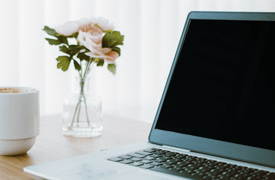 Ein Schreibtisch mit Laptop, Tasse und Blumenvase aus Glas mit Schnittrosen.