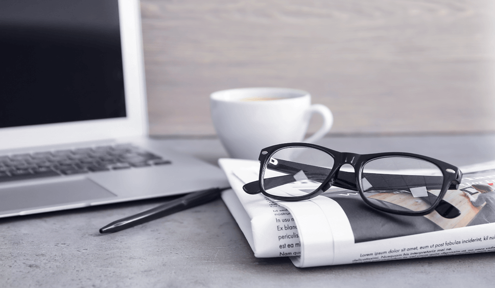 Ein Schreibtisch mit Laptop, Tasse, Kugelschreiber, Zeitung und Brille.