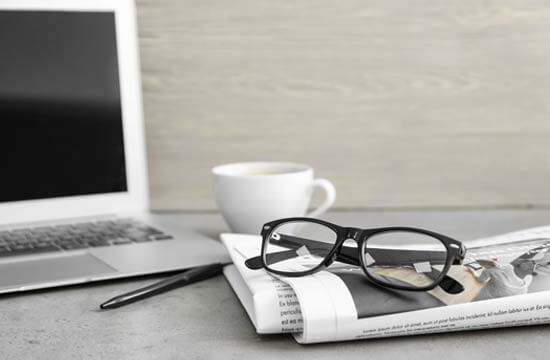 Ein Schreibtisch mit Laptop, Tasse, Kugelschreiber, Zeitung und Brille.