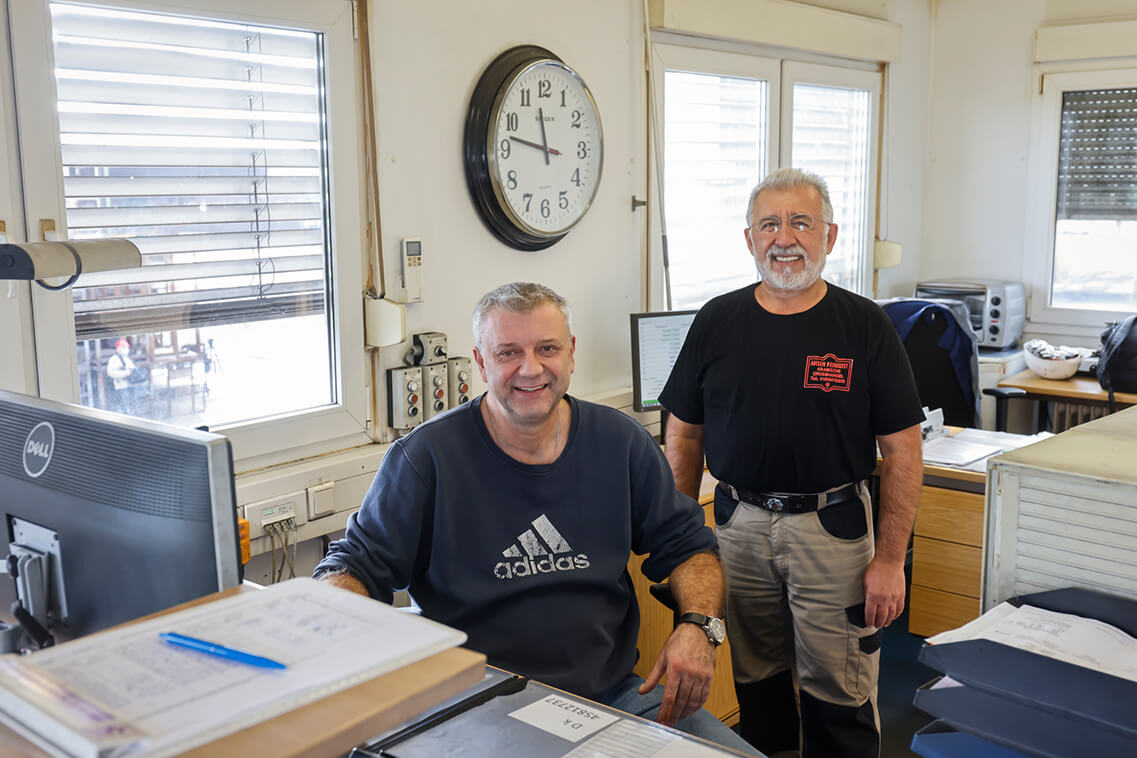 Das Foto wurde im Büro der Waage aufgenommen. Frank Esser trägt Jeans und ein blaues Sweatshirt, er sitzt. Neben ihm steht Asaf Öztürk. Er trägt ein schwarzes T-Shirt und eine beige Arbeitshose. Beide lachen, im Hintergrund ist eine große Uhr zu sehen, auf der es 10 vor zwölf ist.