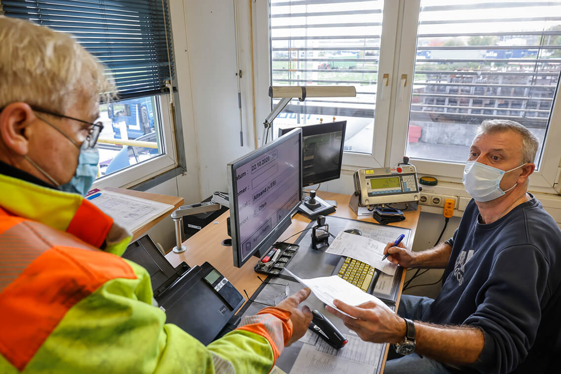  An einem Schreibtisch sitzt Frank Esser vor einem Bildschirm. Vorne in der Tür steht ein Lastwagenfahrer mit Helm und einer Schutzweste und überreicht ihm ein Klemmbrett mit Papieren. Beide abgebildeten Personen tragen eine Mund-Nasen-Bedeckung.