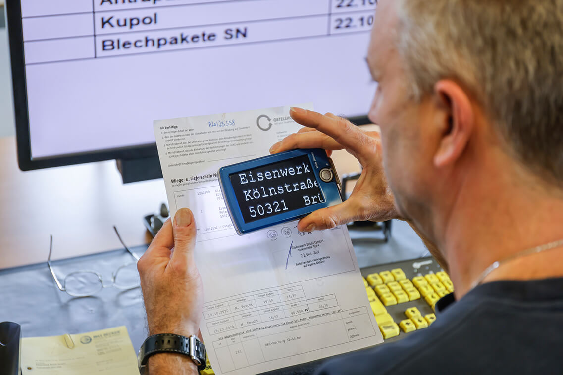 Das Foto ist ein Blick über die Schulter von Frank Esser, der am Schreibtisch sitzt und Frachtpapiere prüft. Er benutzt dafür eine elektronische Lupe, die aktuell auf das Adressfeld des Eisenwerks gerichtet ist. Auf dem Bildschirm der Lupe sieht man stark vergrößert „Eisenwerk“ und „Kölnstraße“. Im Hintergrund auf dem Bildschirm ist die Schrift ebenfalls stark vergrößert.
