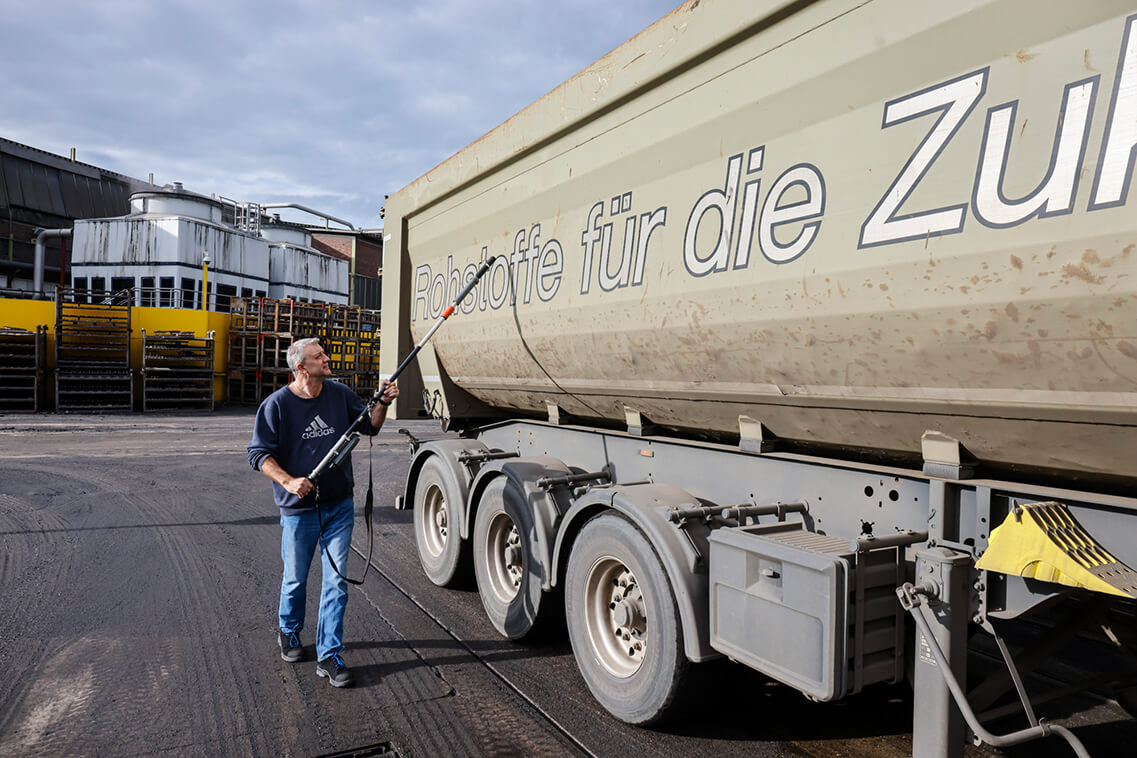 Frank Esser trägt Jeans und ein blaues Sweatshirt und läuft mit einem Strahlenmessgerät an der Ladefläche eines Lasters entlang. Das Gerät ist auf einem Teleskopstab montiert. Der Laster hat eine graugrüne Ladefläche, an der Seite steht in großer Typo „Rohstoffe für die Zukunft“.