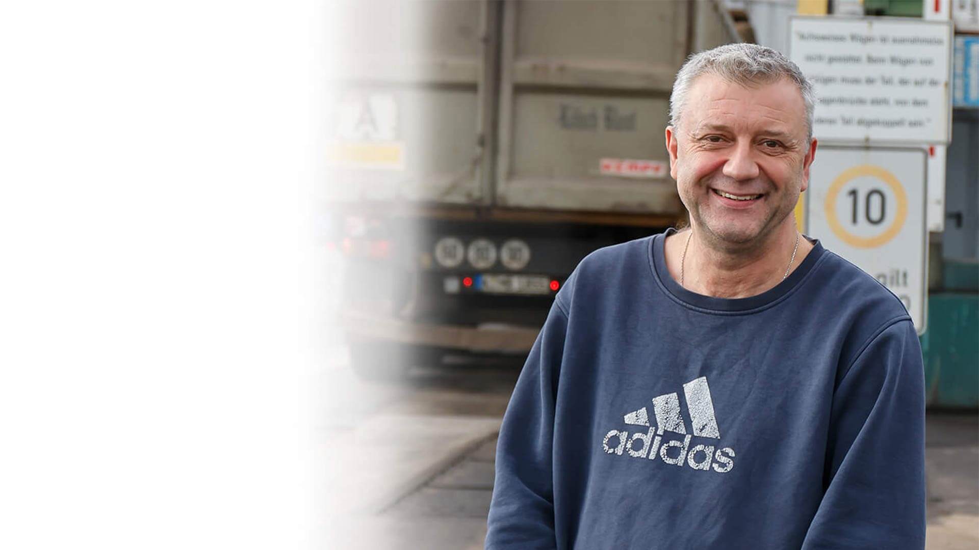 Halbportrait von Frank Esser. im Freien Er trägt ein blaues Sweatshirt und lächelt in die Kamera. In Hintergrund ein LKW.