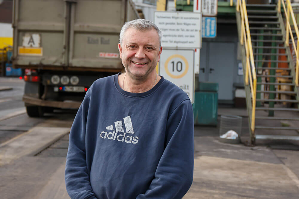 Frank Esser steht an der Werkseinfahrt, im Hintergrund ist ein Lastwagen und ein Containerhäuschen, zu dem eine Treppe mit gelbem Geländer hinaufführt, zu sehen. Frank Esser trägt ein blaues Sweatshirt mit dem Adidas-Logo auf der Brust, sowie Jeans. Er lächelt und hält die Hände vor dem Bauch verschränkt.