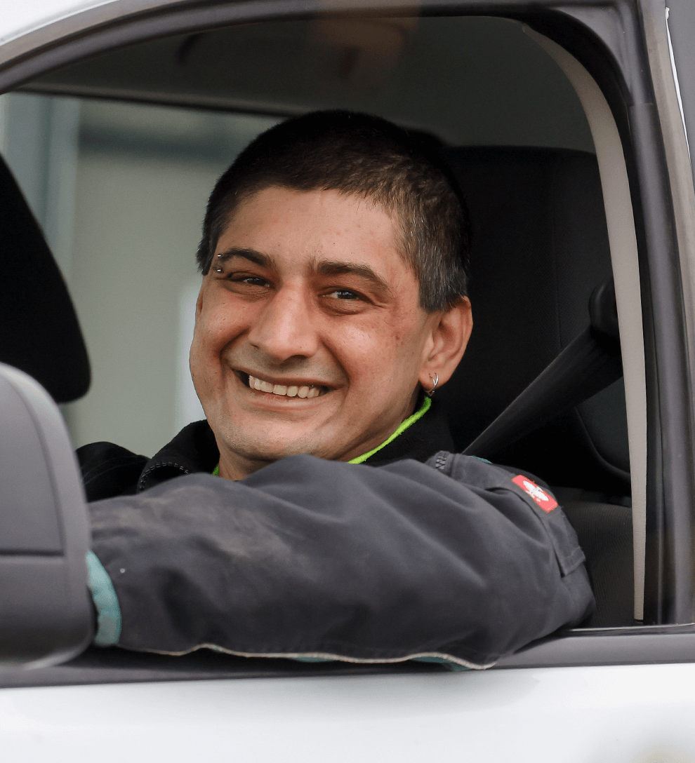 Michael Schmitz sitzt im Auto hinter dem Steuer und hat den Ellbogen aus dem geöffneten Fenster gestreckt. Er trägt eine graue Arbeitsjacke und lächelt freundlich.