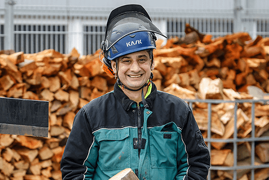 Michael Schmitz ist Mitarbeiter im GaLa-Service Rhein-Erft, einem Garten- und Landschaftsunternehmen. Er steht vor einem Holzstapel und trägt einen blauen Helm und Arbeitskleidung. Michael Schmitz schaut freundlich lächelnd.