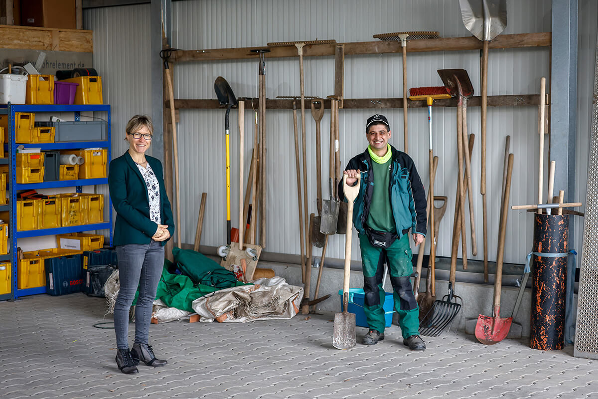 Birgit Hummel und Michael Schmitz stehen im Abstand von zwei Metern in einer Gerätehalle des Garten- und Landschaftsbaubetriebes. An der Rückwand der Halle befinden sich Gartengeräte. Beide lächeln freundlich.