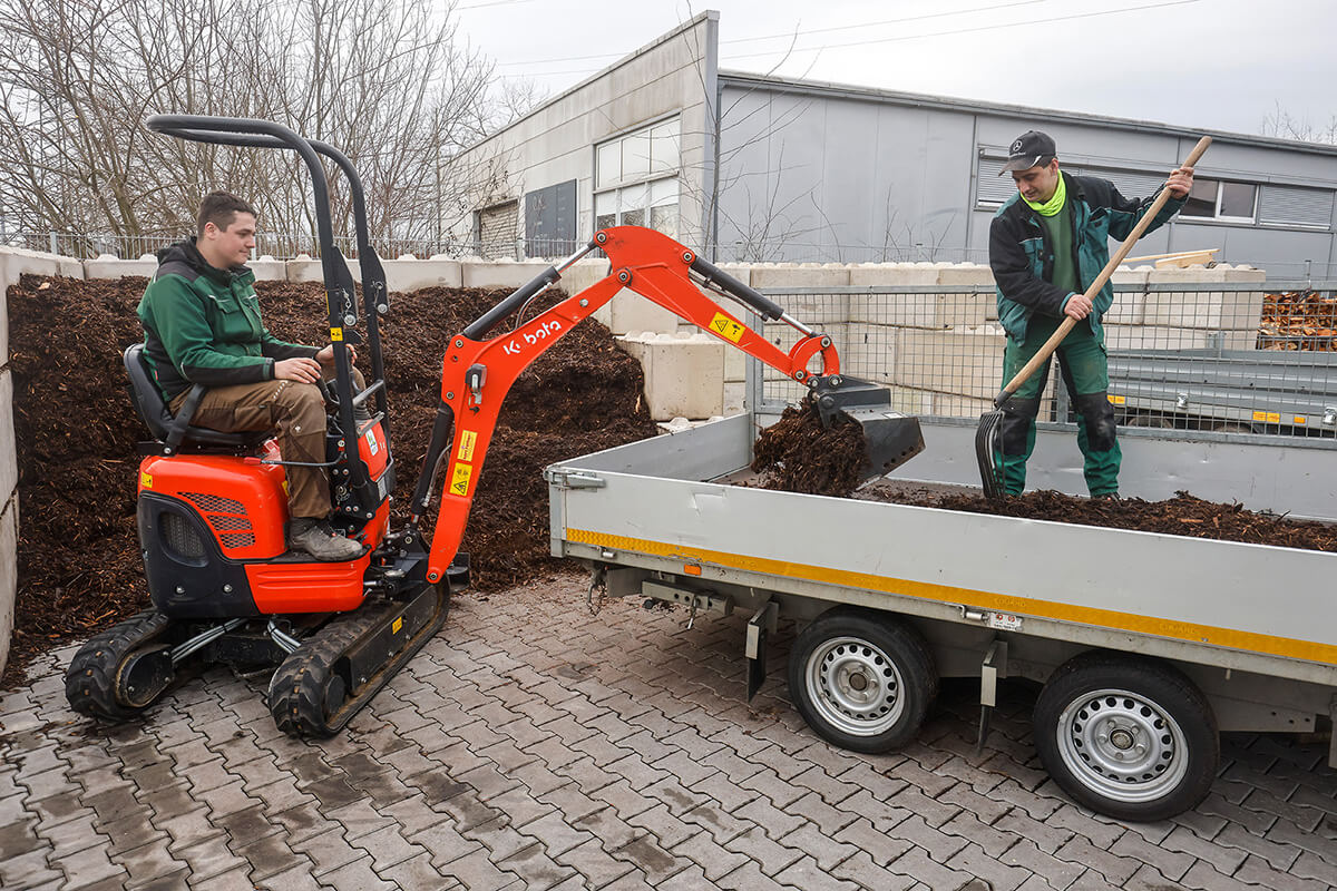 Michael Schmitz und ein Kollege beladen einen Anhänger mit Rindenmulch. Michael Schmitz steht mit einer Harke auf dem Anhänger - der Kollege sitz im Bagger und belädt den Anhänger. Beide tragen Arbeitskleidung.