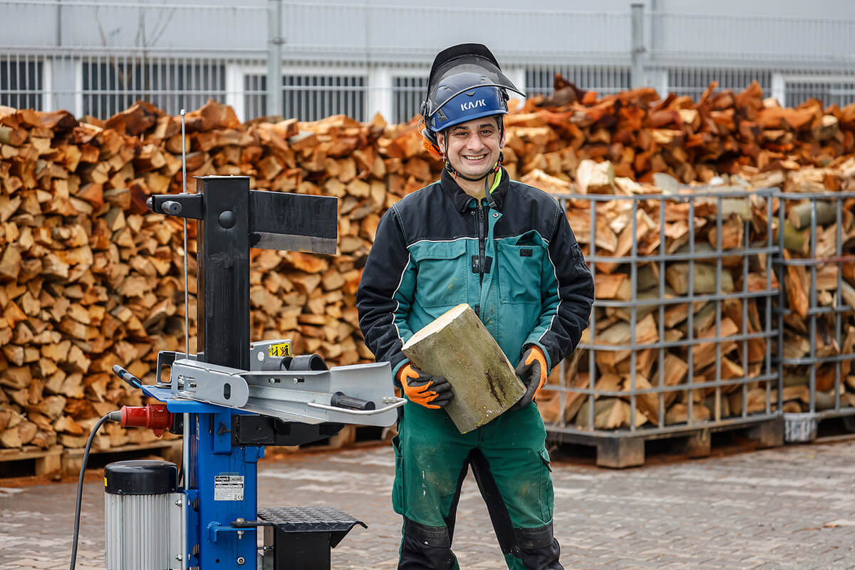 Michael Schmitz ist Mitarbeiter im GaLa-Service Rhein-Erft, einem Garten- und Landschaftsunternehmen. Er steht vor einem Holzstapel und trägt einen blauen Helm und Arbeitskleidung. Michael Schmitz schaut freundlich lächelnd.